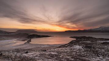 A coastal landscape with a frozen lake, snow-covered terrain, and distant mountains under a sunset sky.