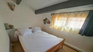 Een slaapkamer met een houten bed, witte beddengoed, een raam met dunne gordijnen en een klein plankje met een decoratief object.