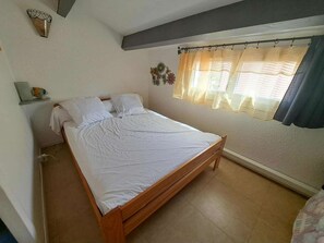 A bedroom with a wooden bed, white bedding, a window with sheer curtains, and a small shelf with a decorative item. - Terraced House with Terrace and Parking - Marseillan-Plage (Marseillan-Plage)