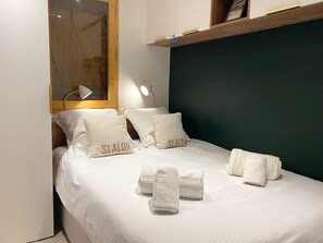 Ein ordentlich bezogenes Bett mit Kissen mit dem Aufdruck "SLALON", ein Fenster mit Jalousien und eine Nachttischlampe.