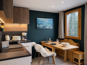 Eine moderne Küche mit Holzschränken, einem Esstisch mit Stühlen, einer Kaffeemaschine und einem Fenster mit Blick auf Bäume.