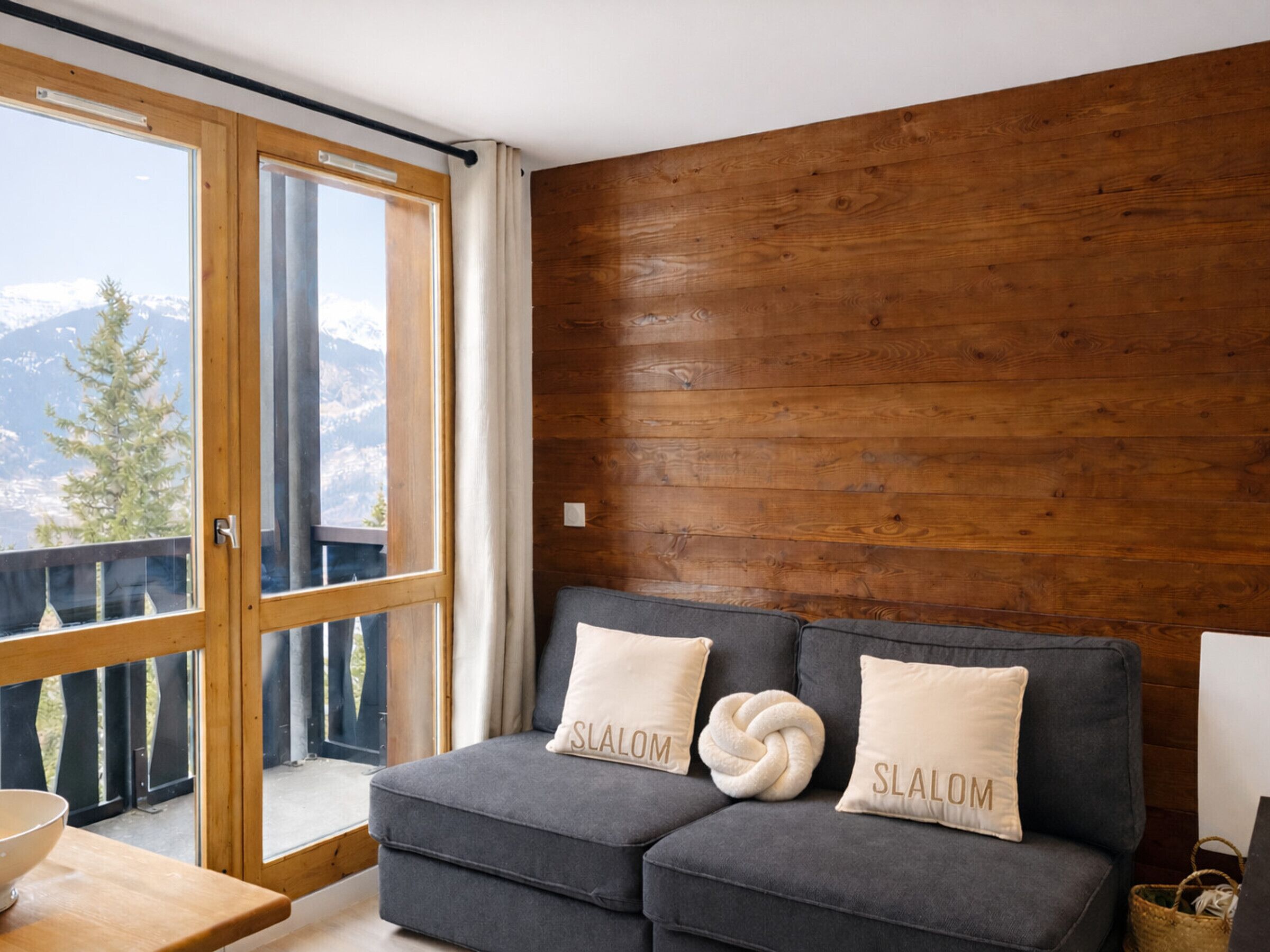Ein gemütliches Zimmer mit einem grauen Sofa, zwei Kissen mit dem Aufdruck "SLALOM", einer Holzwand und Bergblick durch eine Glastür.