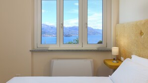 Habitación de hotel con cama, mesita de noche con lámpara y ventana con vista a montañas y agua.