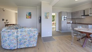 Una sala de estar moderna con un sofá floral, un comedor con una mesa redonda y sillas transparentes, y una cocina con gabinetes grises y un horno empotrado.
