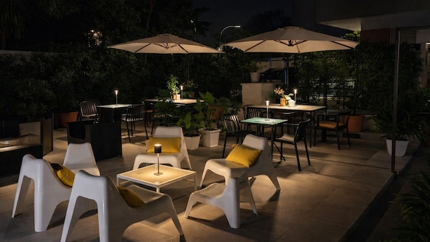 Un patio al aire libre con mobiliario moderno, incluyendo sillas blancas con cojines amarillos, una mesa y dos sombrillas que dan sombra.