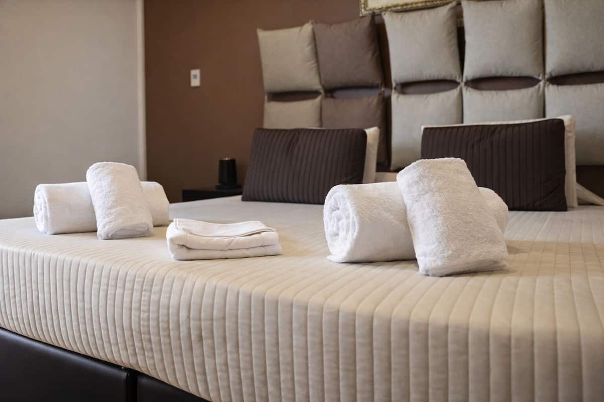A neatly made bed with rolled white towels, a headboard with multiple cushions, and a bedside table with a lamp.