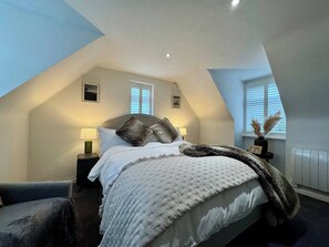 Un dormitorio con techo inclinado, cama con edredón blanco y manta gris, dos lámparas de noche y una ventana con persianas.