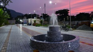 A tiled fountain with water jets, surrounded by a paved walkway, under streetlights, with a mountain in the background. - SRI VIJAYBALAJI MAHAL  (Tiruvannamalai)