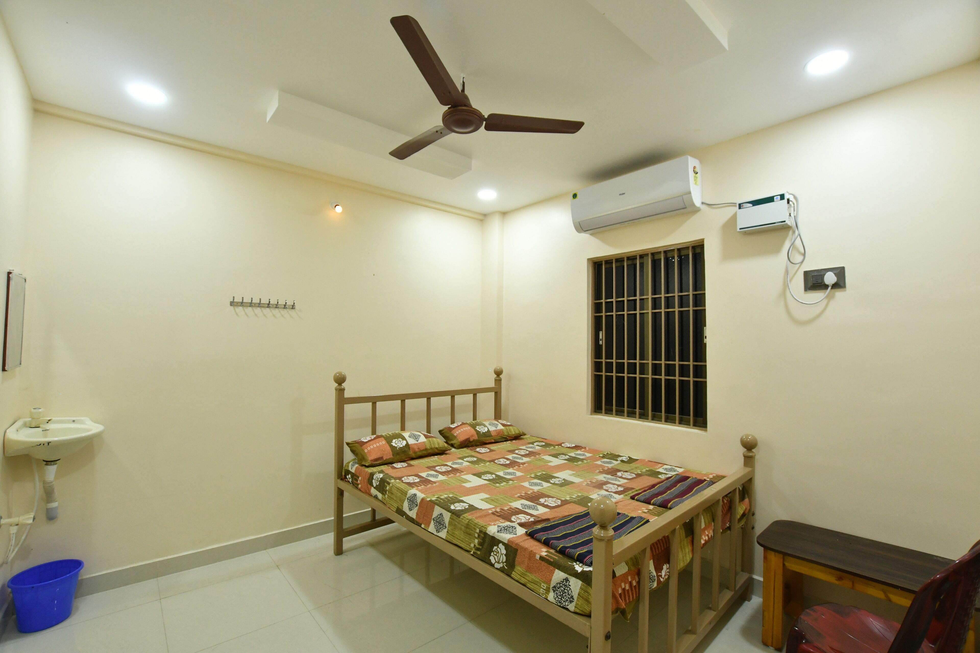 A bedroom with a bed, a ceiling fan, an air conditioning unit, a window with bars, a bedside table, and a small sink.