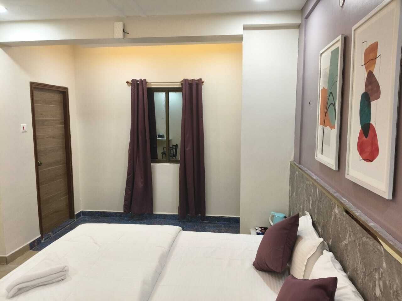 A hotel room with a bed, a door, a window with curtains, and two framed artworks on the wall.