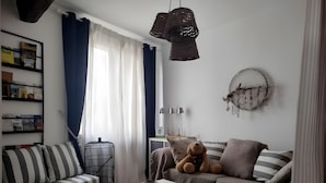 Olohuoneessa on raidallinen sohva, valkoinen sohvapöytä, pehmolelu, kirjahylly ja ikkuna, jossa on verhot.