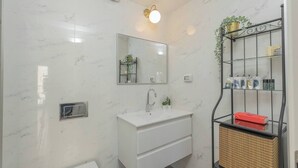A bathroom with a white marble wall, a mirror, a sink, a towel rack, and a shelf with toiletries. - 1 BR Apartment in Ha-Neviim (w/ safe room) (Jerusalem)