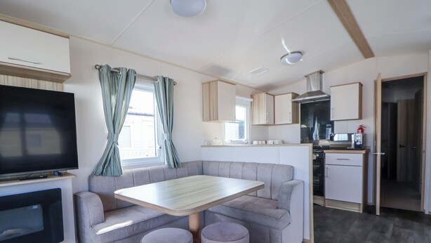 A compact living space with a sofa, a small table, two ottomans, a TV, and a kitchenette. - The Beachside Hideout - St Michaels Caravan Park (Abergele)