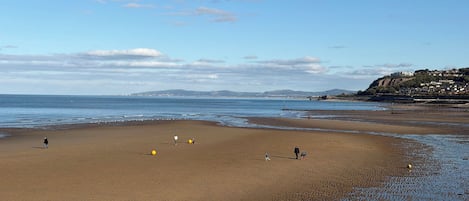 A wide sandy beach with gentle waves, a clear sky, and distant hills.