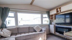 A living room with a sofa, a television, and a window with curtains. - The Beachside Hideout - St Michaels Caravan Park (Abergele)