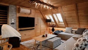 Une pièce confortable, aménagée comme un grenier, avec un plafond en bois, un téléviseur à écran plat, un canapé, une table basse et une petite table d’appoint sur laquelle est posé un vase.