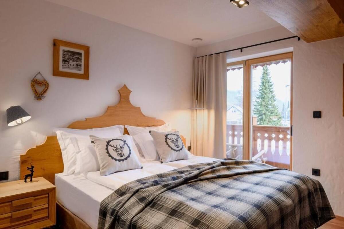 A bedroom with a wooden headboard, a bed with white bedding and a plaid blanket, a nightstand with a lamp, and a window with a view of trees and a house.