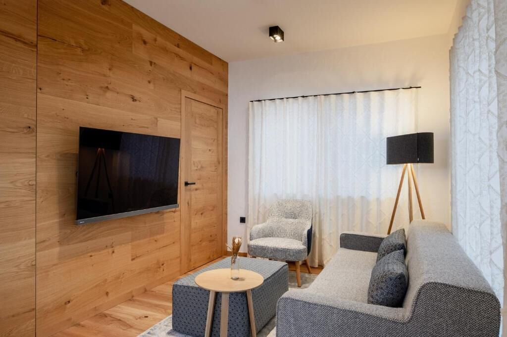A modern living room with a wooden wall, a flat-screen TV, a grey sofa, a patterned armchair, a small round table, and a floor lamp.