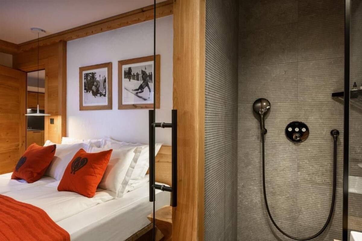A modern hotel room with a wooden ceiling, a bed with white linens and orange pillows, a shower with a control panel, and two framed pictures on the wall.