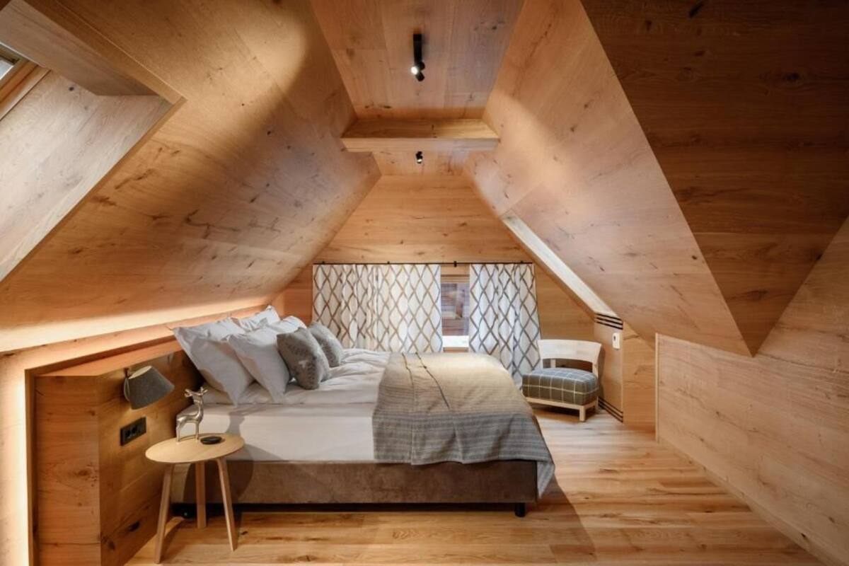 A cozy attic bedroom with a wooden sloped ceiling, a bed with pillows, a small table, and a chair.