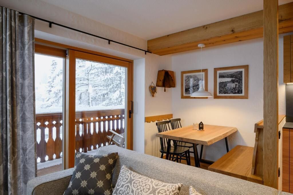 A cozy room with a wooden dining table, a sofa with patterned cushions, and a view of snowy trees through large windows.