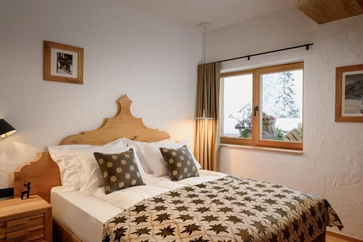 A bedroom with a wooden headboard, a bed with white linens and patterned pillows, a nightstand with a lamp, and a window with curtains.