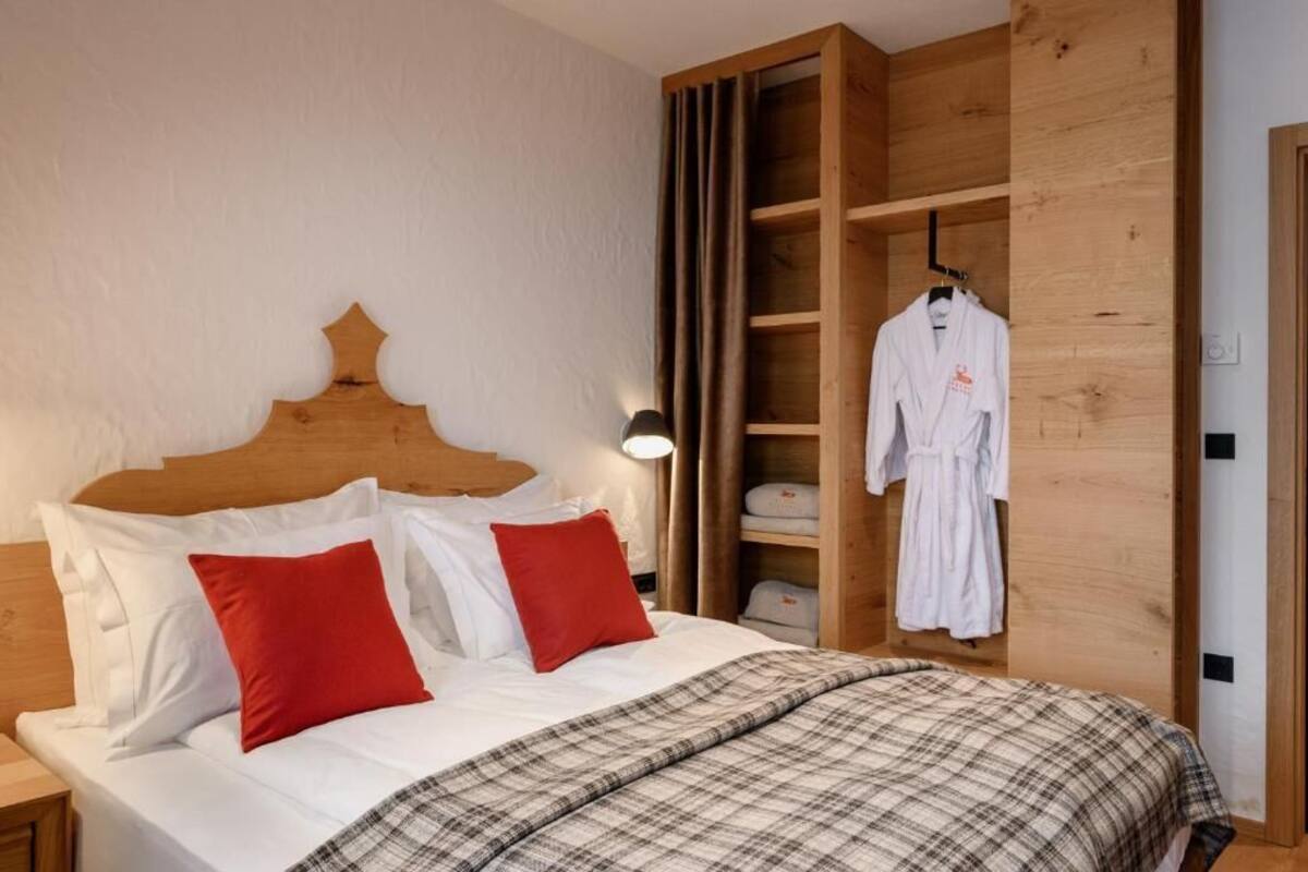 A bedroom with a wooden headboard, a bed with white and red bedding, a nightstand, a bedside lamp, and a wooden wardrobe with a robe hanging.