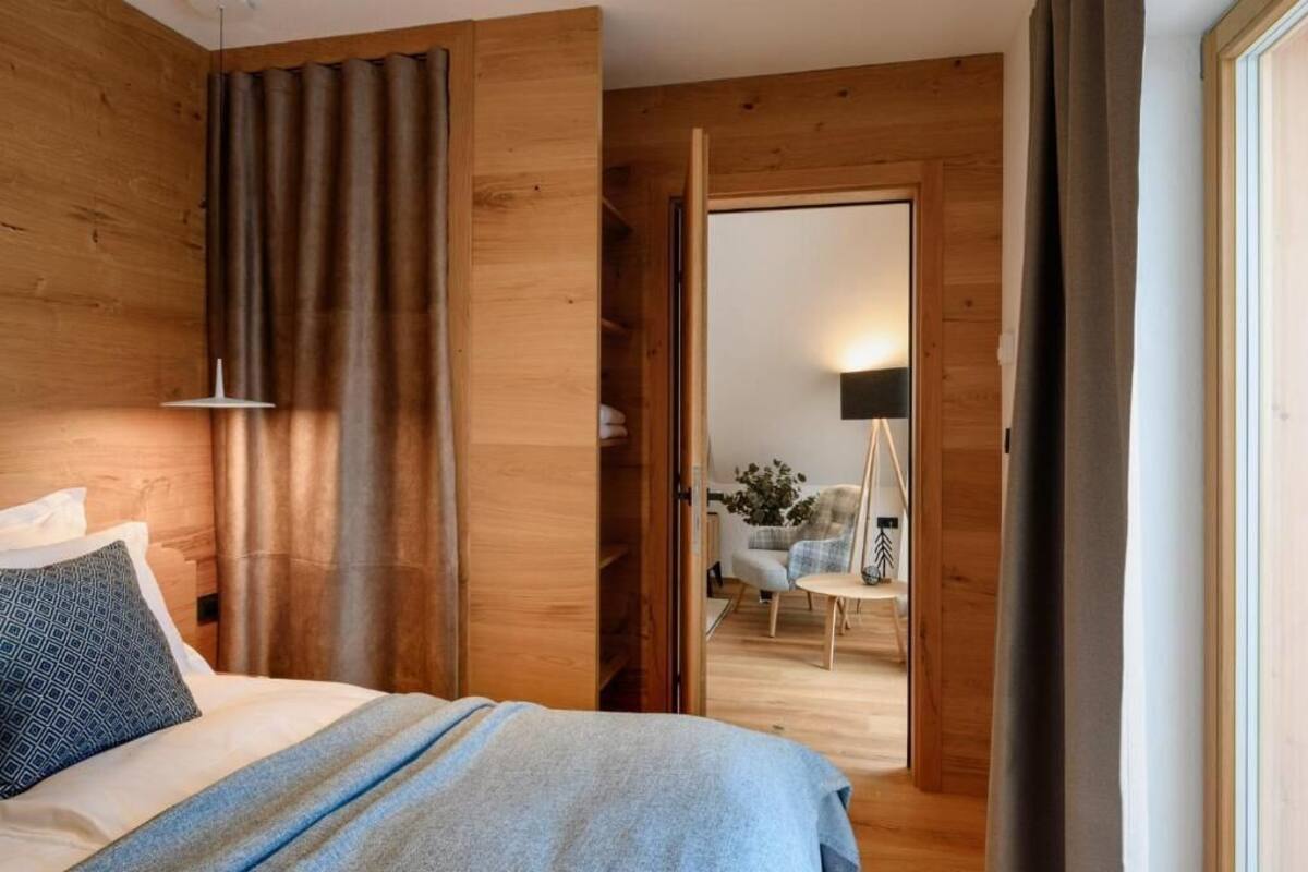 A modern bedroom with wooden walls, a bed with a blue blanket, a bedside lamp, and a view into a dining area with a chair and table.