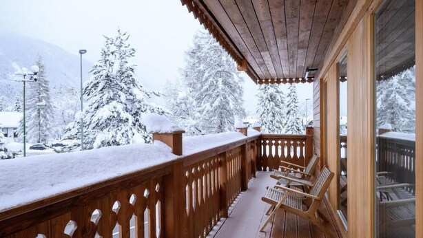 Une terrasse en bois avec une balustrade, deux chaises en bois et une porte vitrée donnant sur un paysage enneigé, avec des arbres et des montagnes en arrière-plan.