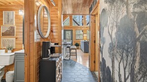 Interior de una cabaña de madera con cocina, comedor y un amplio ventanal con vistas a árboles.