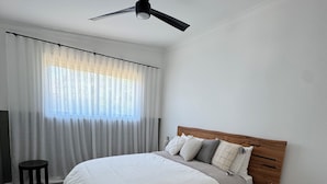 Ein Schlafzimmer mit einem Bett, einem Nachttisch aus Holz, einem Deckenventilator und einem Fenster mit Vorhängen.