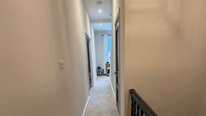 Un estrecho pasillo alfombrado con barandilla de madera en un lado, que conduce a una habitación al final, con una ventana y una puerta.