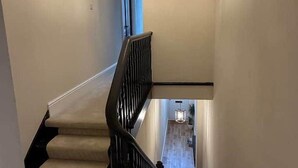 Una escalera estrecha con barandilla negra y alfombra color crema, que conduce a una habitación con suelo de madera y una lámpara colgante.