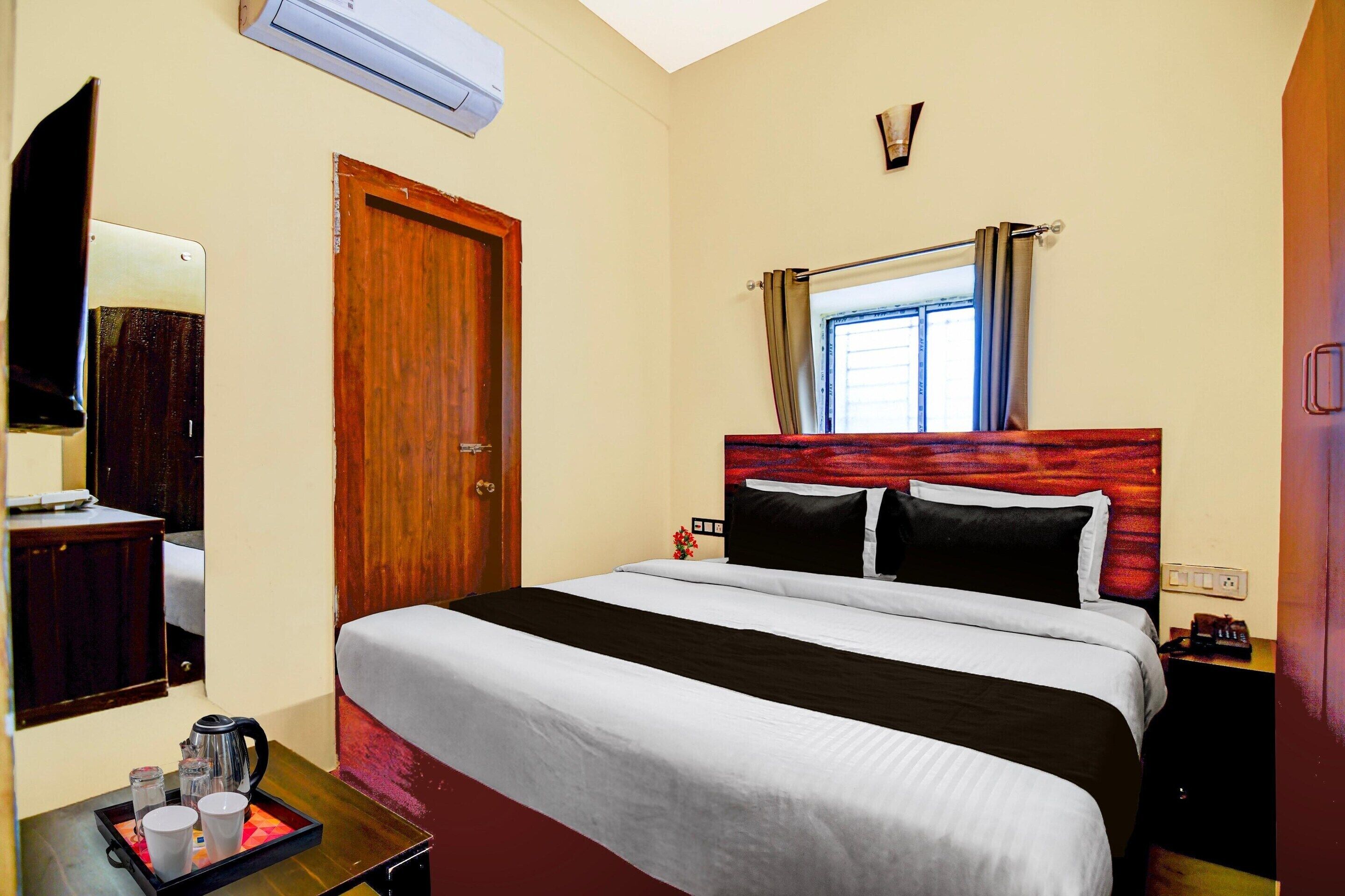 A hotel room with a bed, a bedside table with a tray of items, a window with curtains, a wall-mounted TV, and a door.