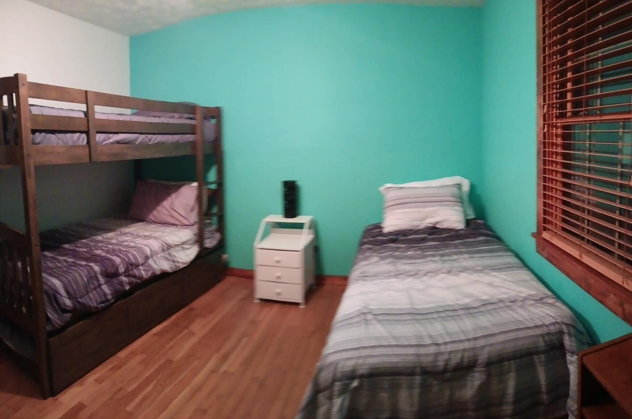A room with a bunk bed, a single bed, a bedside table, and a window with blinds.