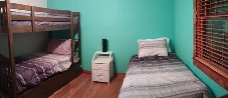 A room with a bunk bed, a single bed, a bedside table, and a window with blinds.