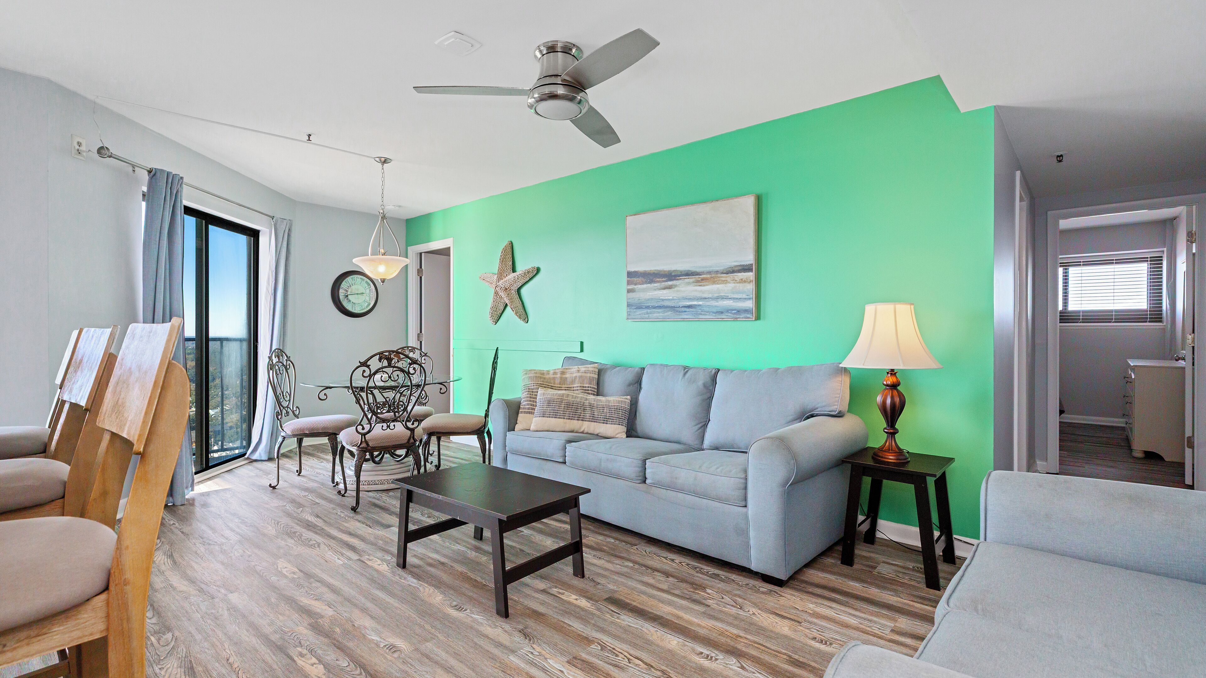 A living room with a green wall, a ceiling fan, a dining table with chairs, a sofa, a lamp, and a starfish decoration.