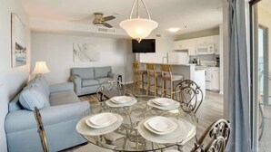 A modern living room with a glass dining table set for four, a sofa, and a TV mounted on the wall. - Penthouse 2BR, Wi-Fi, SMART TVs, Amazing Views! (Myrtle Beach)