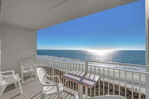 A balcony with white plastic chairs and a small table, overlooking a beach with the ocean and a clear sky. - Corner Oceanview Condo w/ Balcony + Pool (North Myrtle Beach)