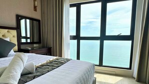 Habitación de hotel con un ventanal que ofrece vistas al mar, una cama bien hecha con almohada y manta, una mesita de noche con lámpara y un espejo en la pared.