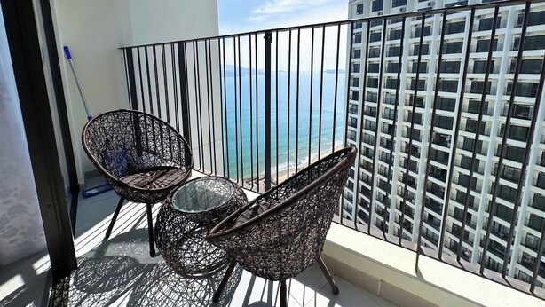 Un balcón con sillas de mimbre y una mesa de vidrio, con vistas a un edificio alto y una masa de agua.