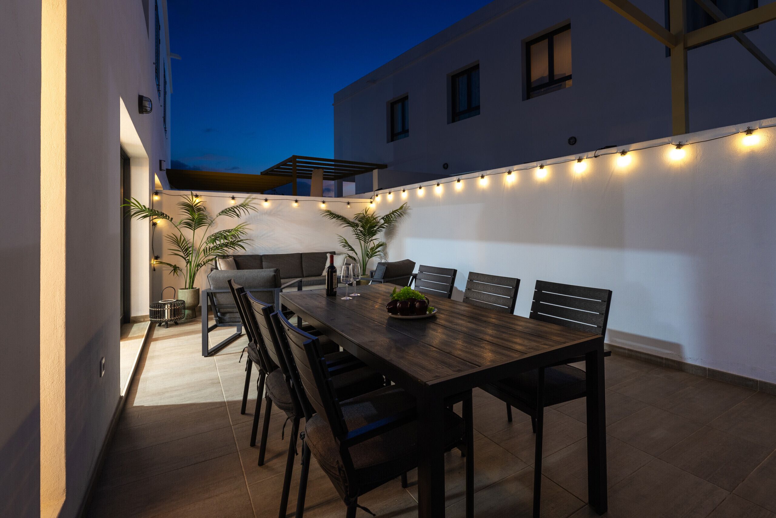 An outdoor dining area with a wooden table and chairs, string lights, and potted plants.