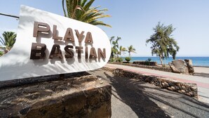 En skylt med texten ”PLAYA BASTIAN”, placerad framför en bakgrund av en strand och palmer.