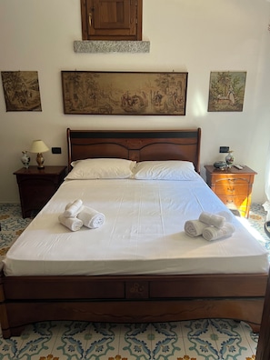 Uma cama bem arrumada com lençóis e toalhas brancos, uma cabeceira de madeira e uma mesinha de cabeceira com um abajur e um pequeno vaso.