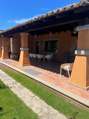 A covered outdoor dining area with a tiled floor, white chairs, and a stone wall. - Vacation Home (Sardegna)
