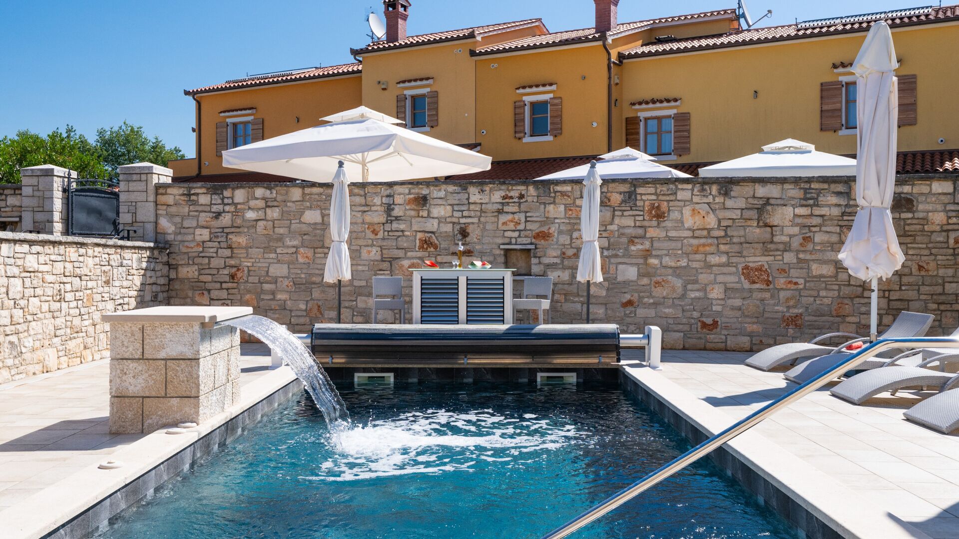 A pool with a water feature, lounge chairs, umbrellas, and a stone wall.