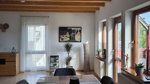 A dining area with a wooden table, chairs, and a floor lamp. There is a window with curtains, a radiator, and a plant on the windowsill. - Spacious vacation apartment (90 sqm) to feel good on 2 levels with balcony (Michelstadt)