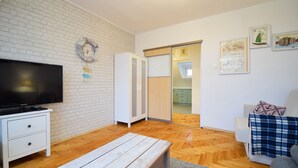 A living room with a flat-screen TV, a white cabinet, a wooden bench, a sofa with a plaid blanket, and a wall with framed artwork. - RentPlanet - Apartament Narutowicza (Kołobrzeg)