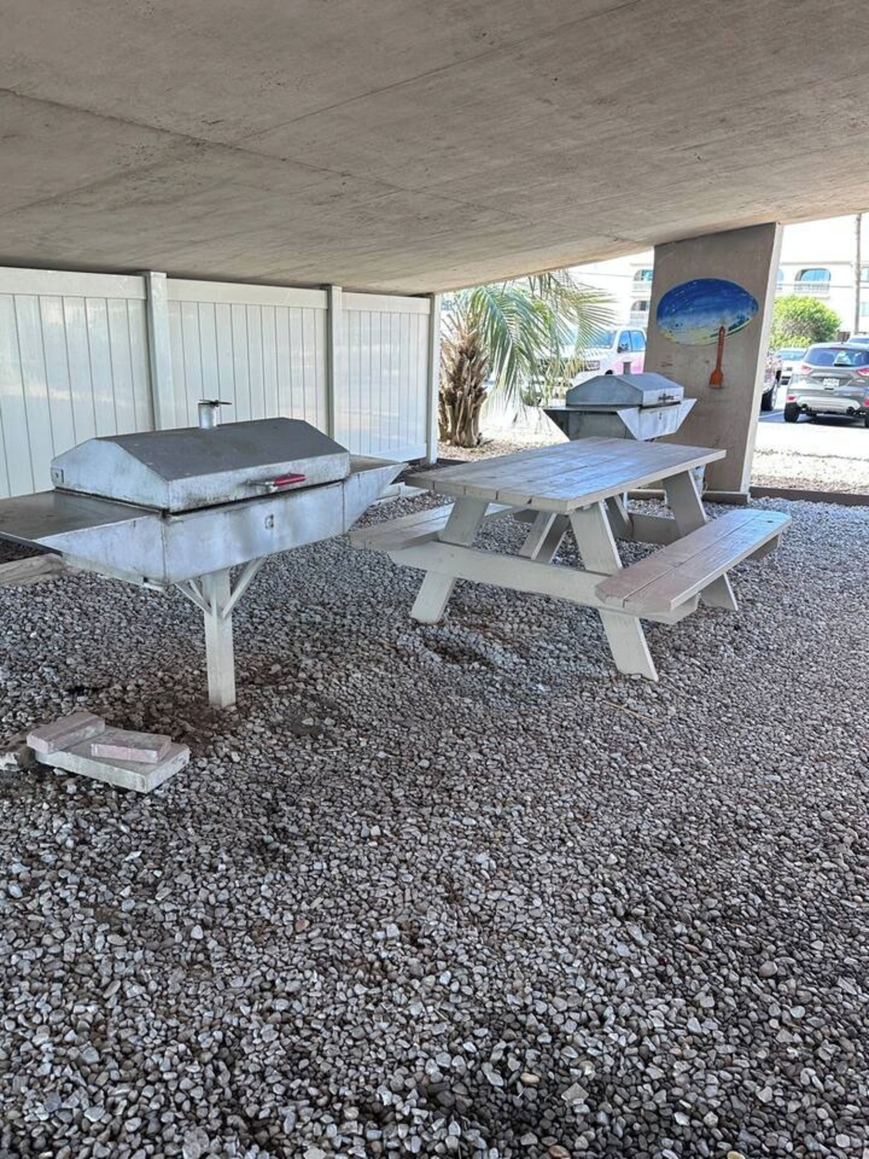 A barbecue grill and picnic table set up on a gravel surface under a covered area.