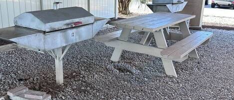 A barbecue grill and picnic table set up on a gravel surface under a covered area.